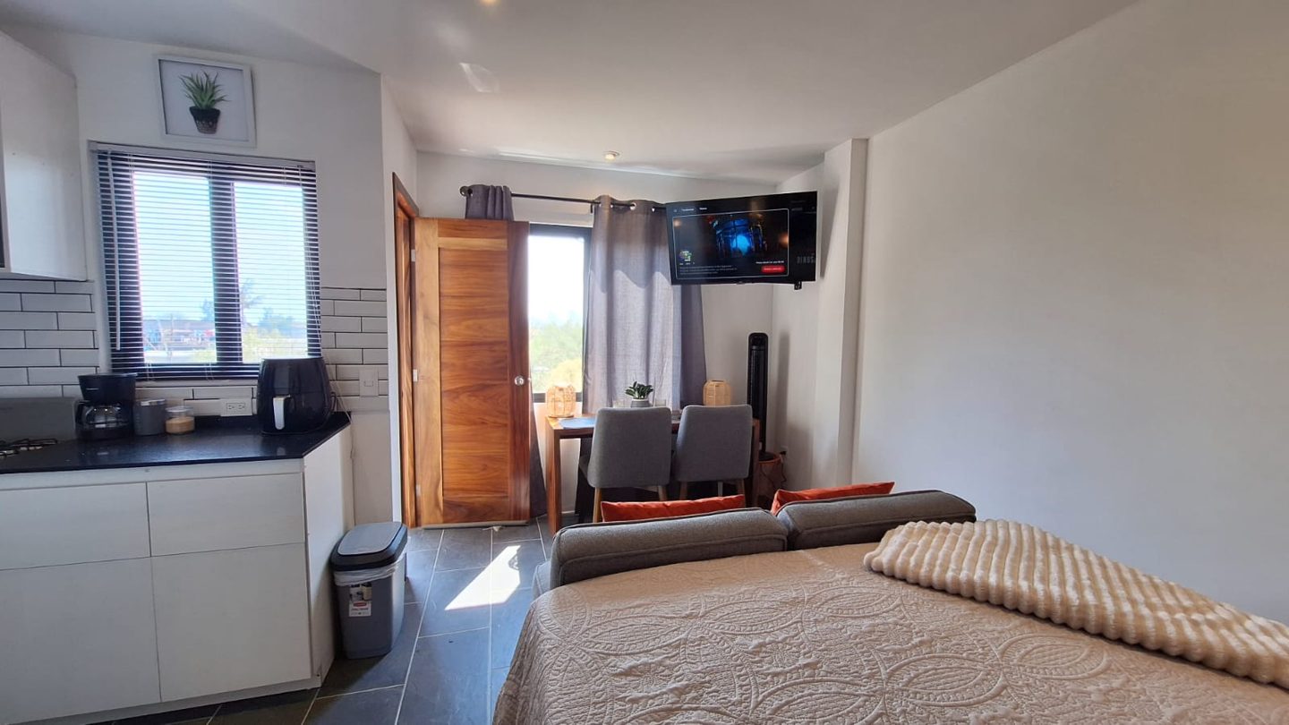 El estudio completo desde la cama: cocina, mesa de comedor, smart TV y la puerta al balcón